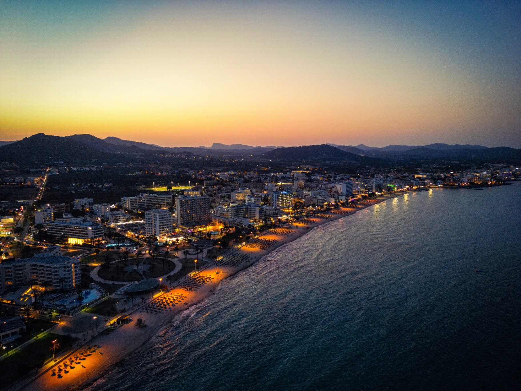 Luftaufnahme der mallorquinischen Küste mit beleuchteter Strandpromenade bei Sonnenuntergang